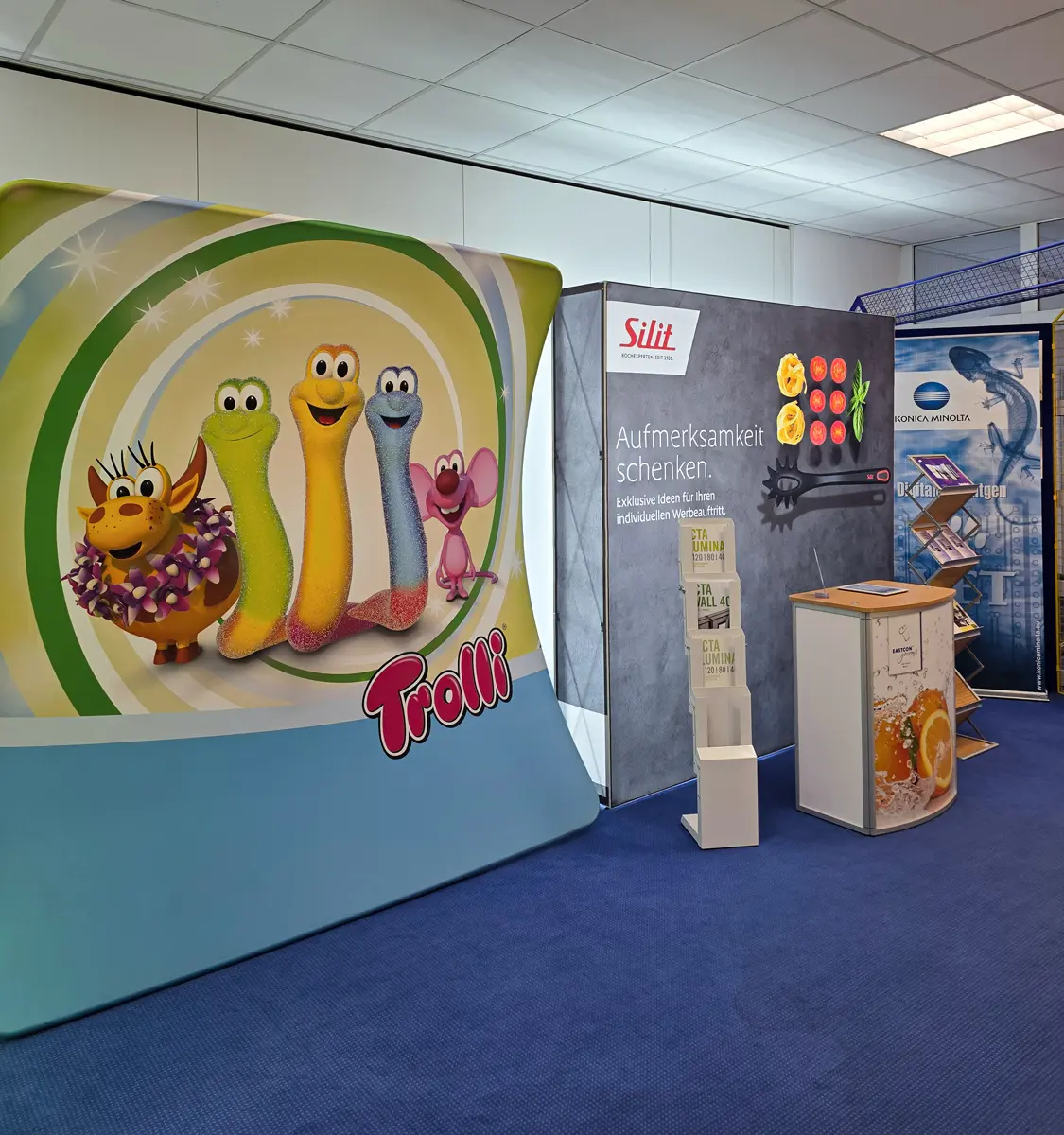Messestand-Displays von Trolli, Silit und Konica Minolta auf blauem Teppich.